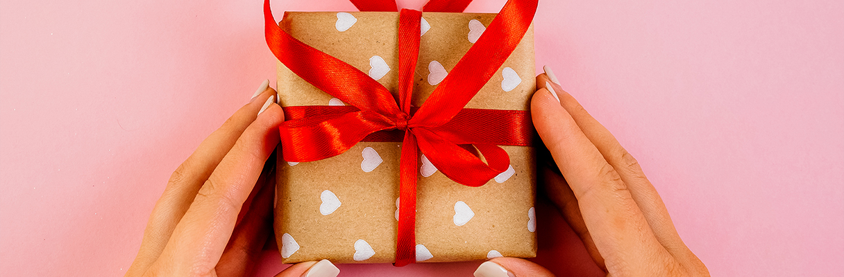Hands holding a small gift wrapped in heart-patterned paper with red ribbon, perfect for romantic anniversaries or special occasions