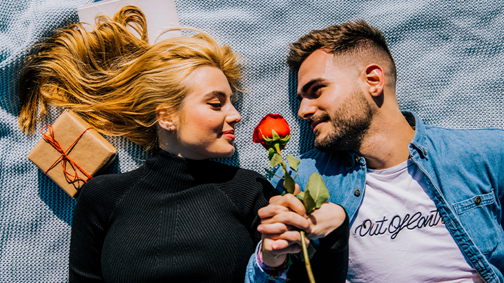 Couple lying on a picnic rug holding hands with a rose and gift, perfect romantic anniversary or special occasion experience
