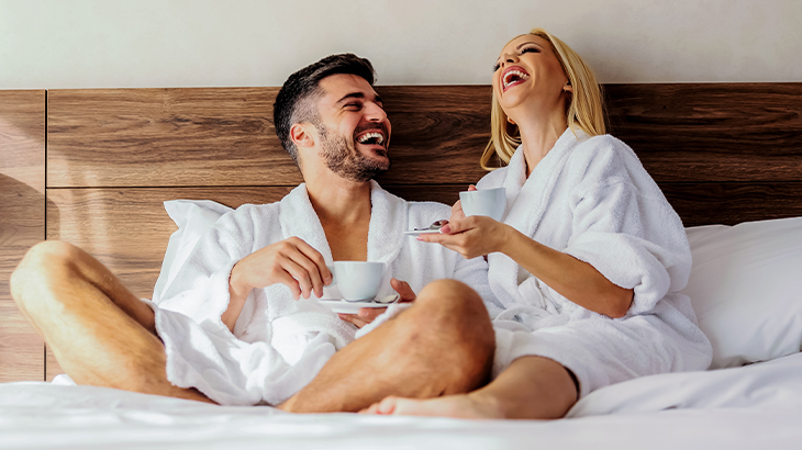 Couple laughing together in robes on a hotel bed during a romantic getaway gift experience, perfect for anniversaries and special occasions