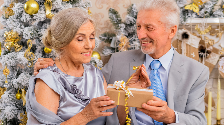 Smartly dressed senior couple exchanging a gift in front of golden Christmas tree and decorations during 50th wedding anniversary holiday celebration