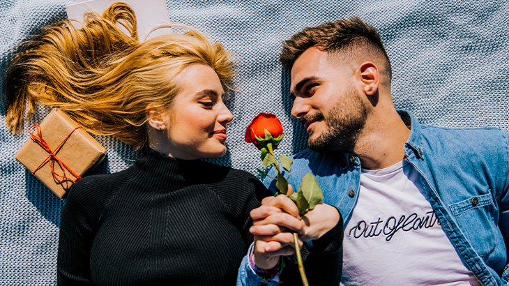 Couple lying on a picnic rug holding hands with a rose and gift, perfect romantic anniversary or special occasion experience