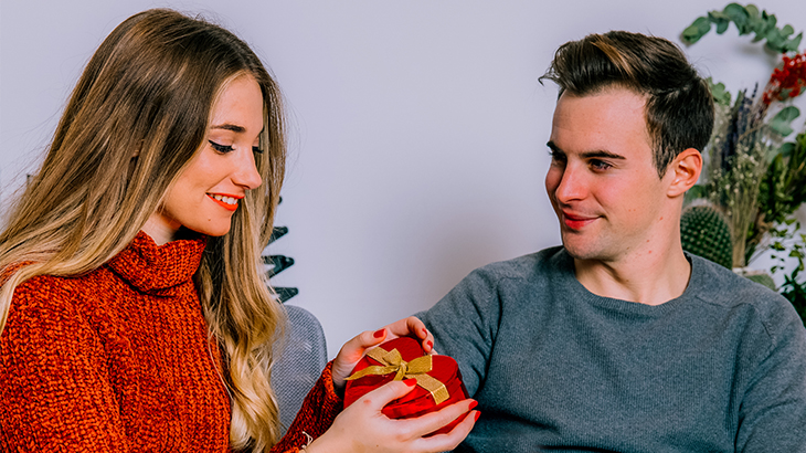 Wife opening a heart-shaped gift from husband while he looks at her lovingly, perfect for anniversary or romantic celebrations