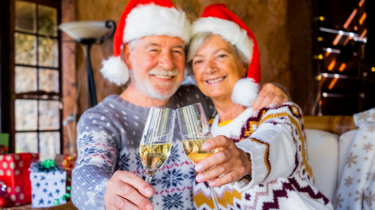 Senior couple in Christmas hats and festive jumpers holding champagne glasses to the camera celebrating 50th wedding anniversary during holiday season