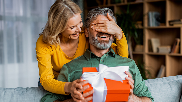 Wife playfully covering husband’s eyes and giving him a 30th wedding anniversary gift to celebrate three decades of love