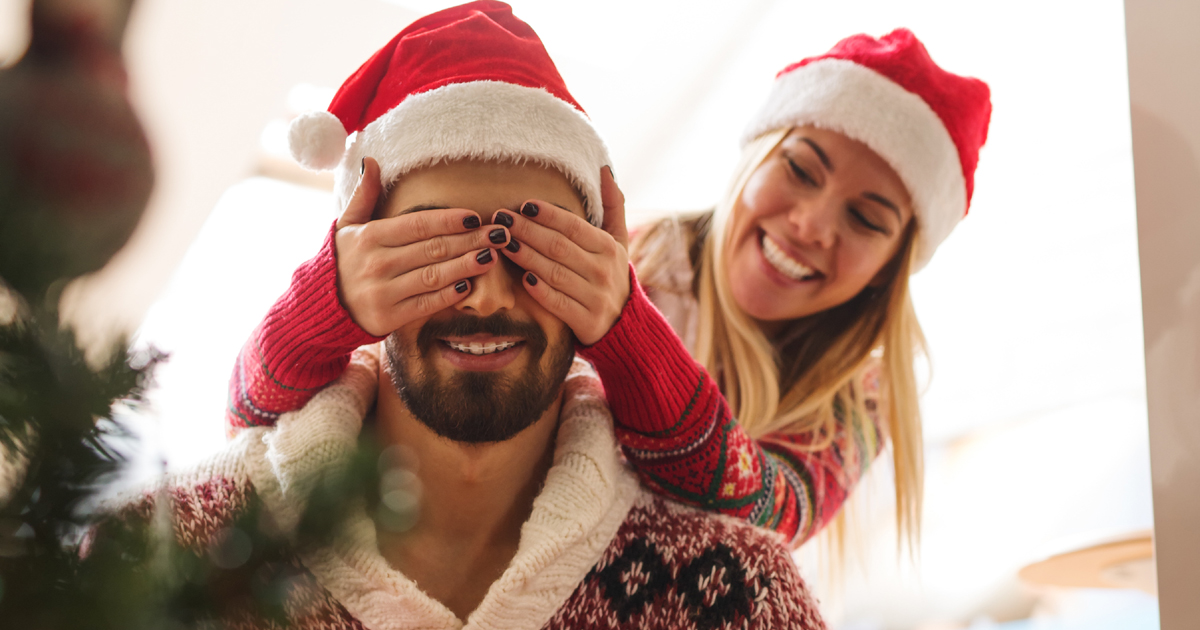 A woman covering her partner's eyes by a Christmas tree A woman covering her partner's eyes by a Christmas tree